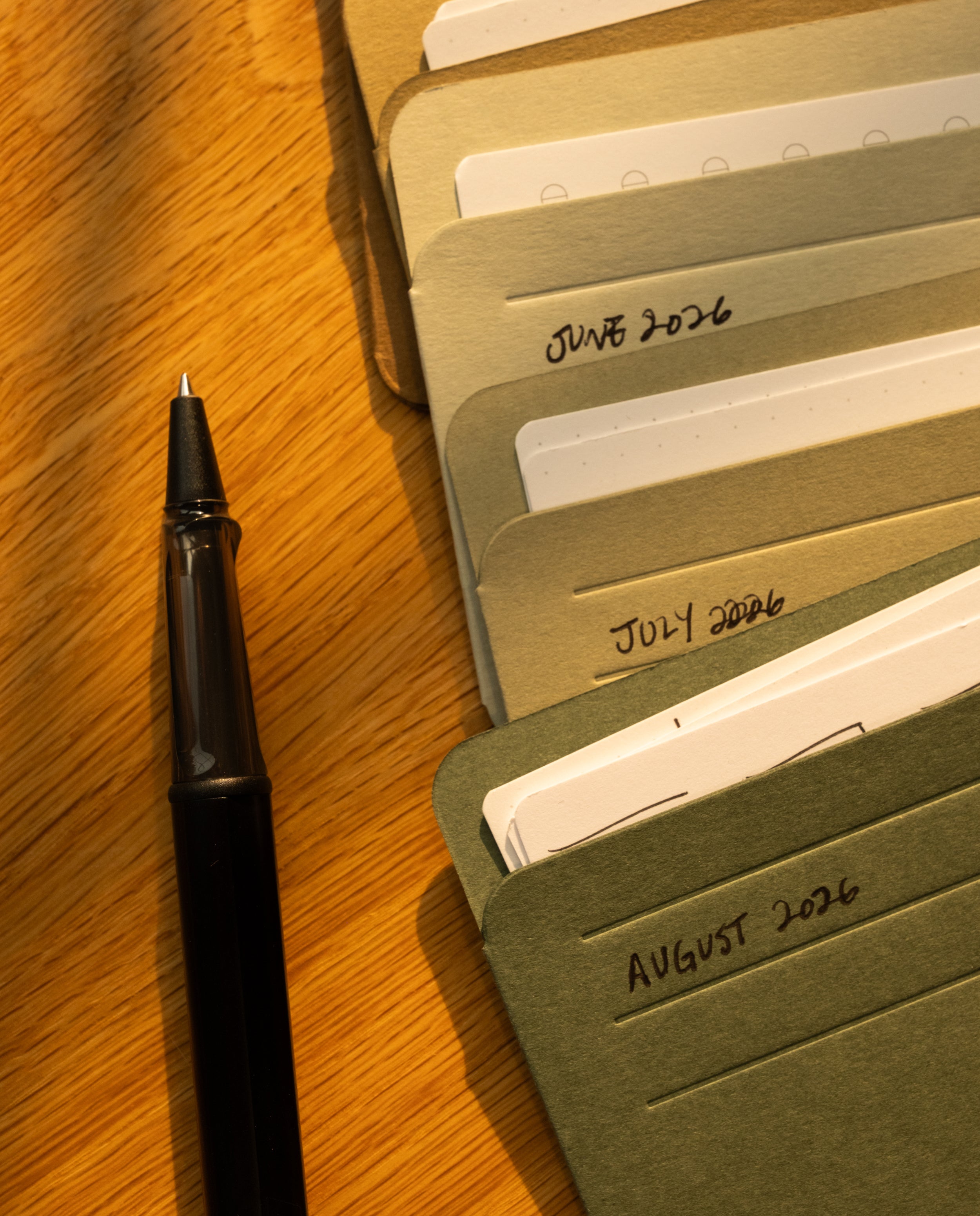 A black pen rests beside several folders labeled June 2026, July 2026, and August 2026, all neatly arranged on a wooden surface under warm lighting. Nearby, an organizer with Analog Card Sleeves - Small (Earth) keeps important notes accessible and protected.