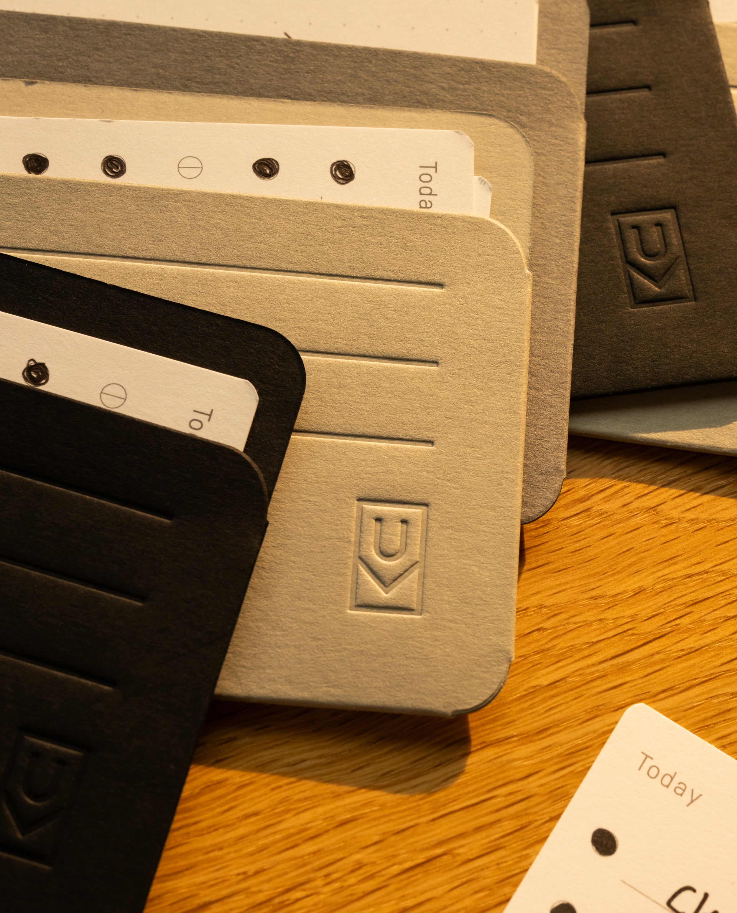 A close-up of black and beige notebooks with embossed logos and lined covers, partially covering sheets of paper labeled “Today,” all on a wooden surface—perfectly complemented by Analog Card Sleeves - Small (Monochrome) for keeping your card organizer neat and accessible.
