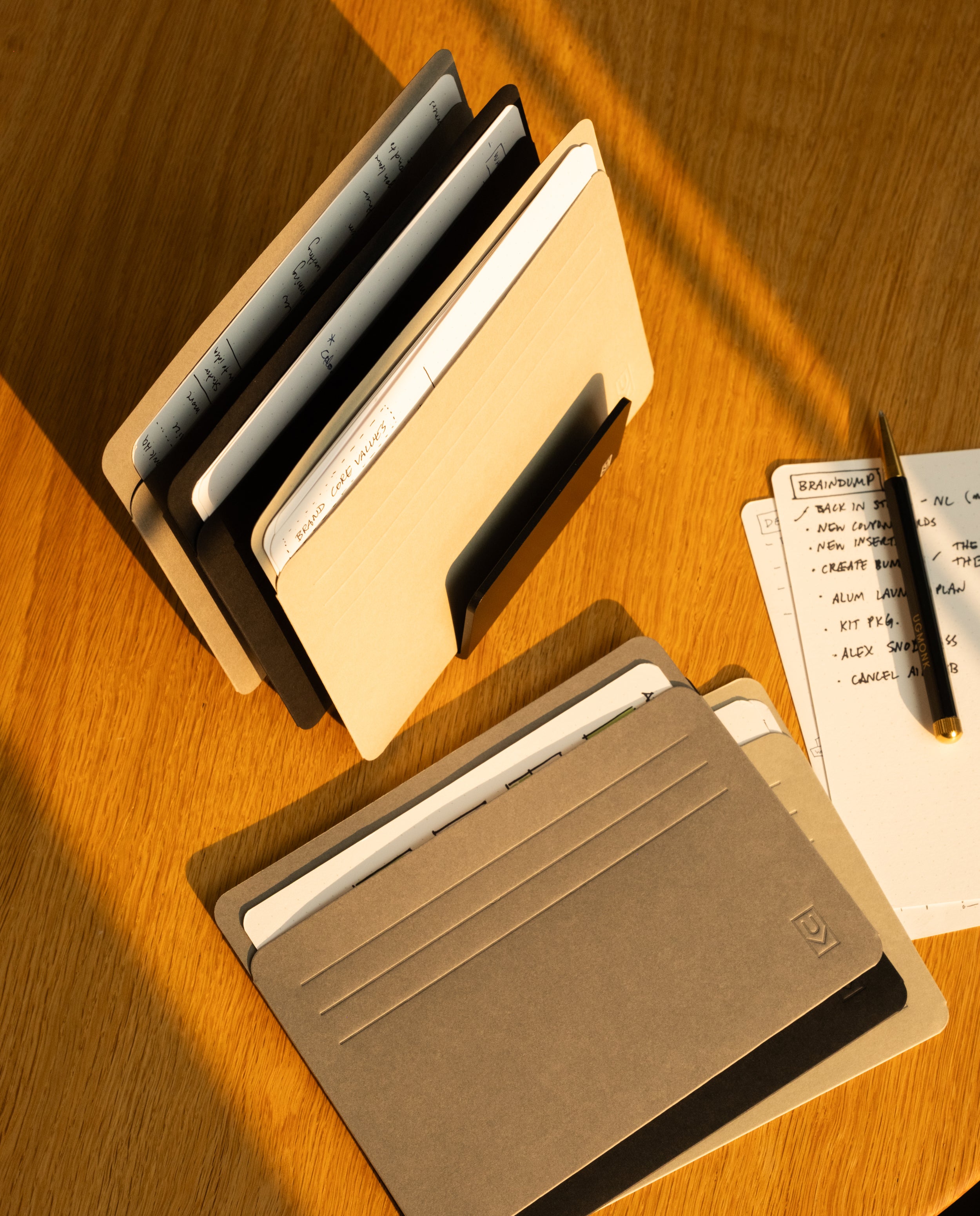 Two sets of notebooks and folders sit on a wooden desk in the warm sunlight. A pen rests on handwritten notes beside them, while a card organizer with Analog Card Sleeves - Large (Monochromatic) enhances the sense of organization and planning in the scene.