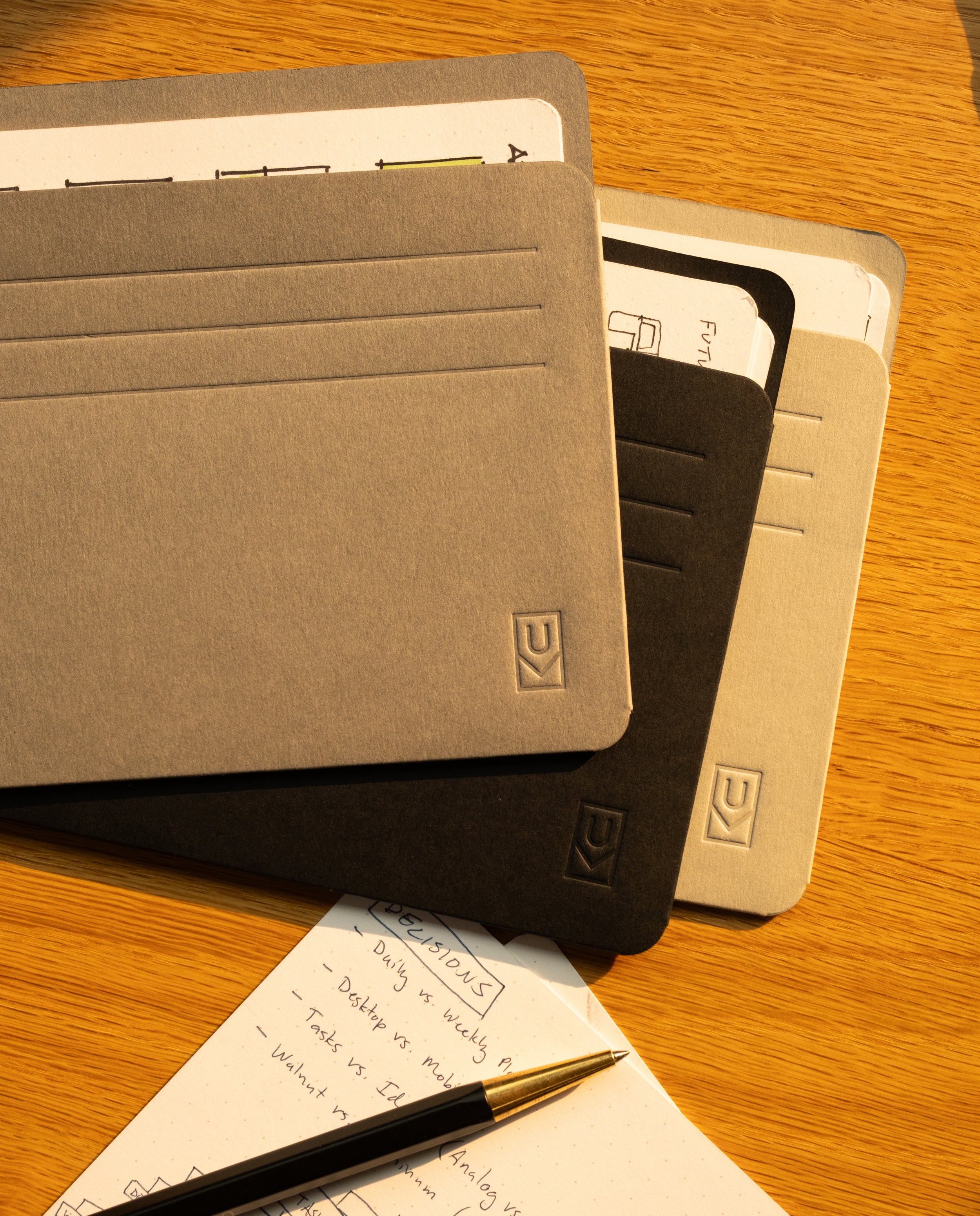 Three notebooks with gray and black covers are stacked on a wooden desk. Nearby, a pen rests on an open notepad filled with handwritten notes and diagrams. Next to them sits the Analog Card Sleeves - Large (Monochromatic) organizer, providing easy access to cards as sunlight casts warm shadows across the scene.