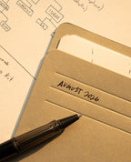 A black pen rests on a folder labeled August 2026, with papers and part of a flowchart or diagram visible beneath it on a dotted sheet, alongside an Analog Card Sleeves - Small (Monochrome) organizer to keep important notes protected and neatly arranged.