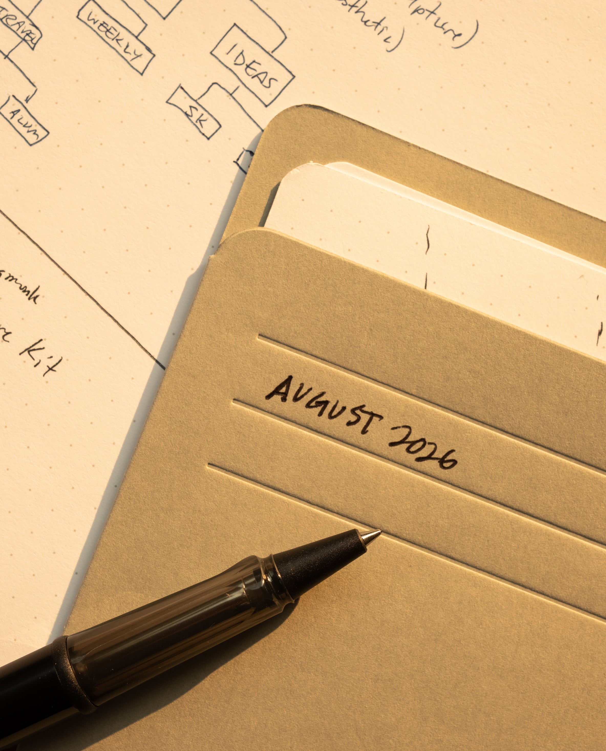 A black pen rests on a folder labeled August 2026, with papers and part of a flowchart or diagram visible beneath it on a dotted sheet, alongside an Analog Card Sleeves - Small (Monochrome) organizer to keep important notes protected and neatly arranged.