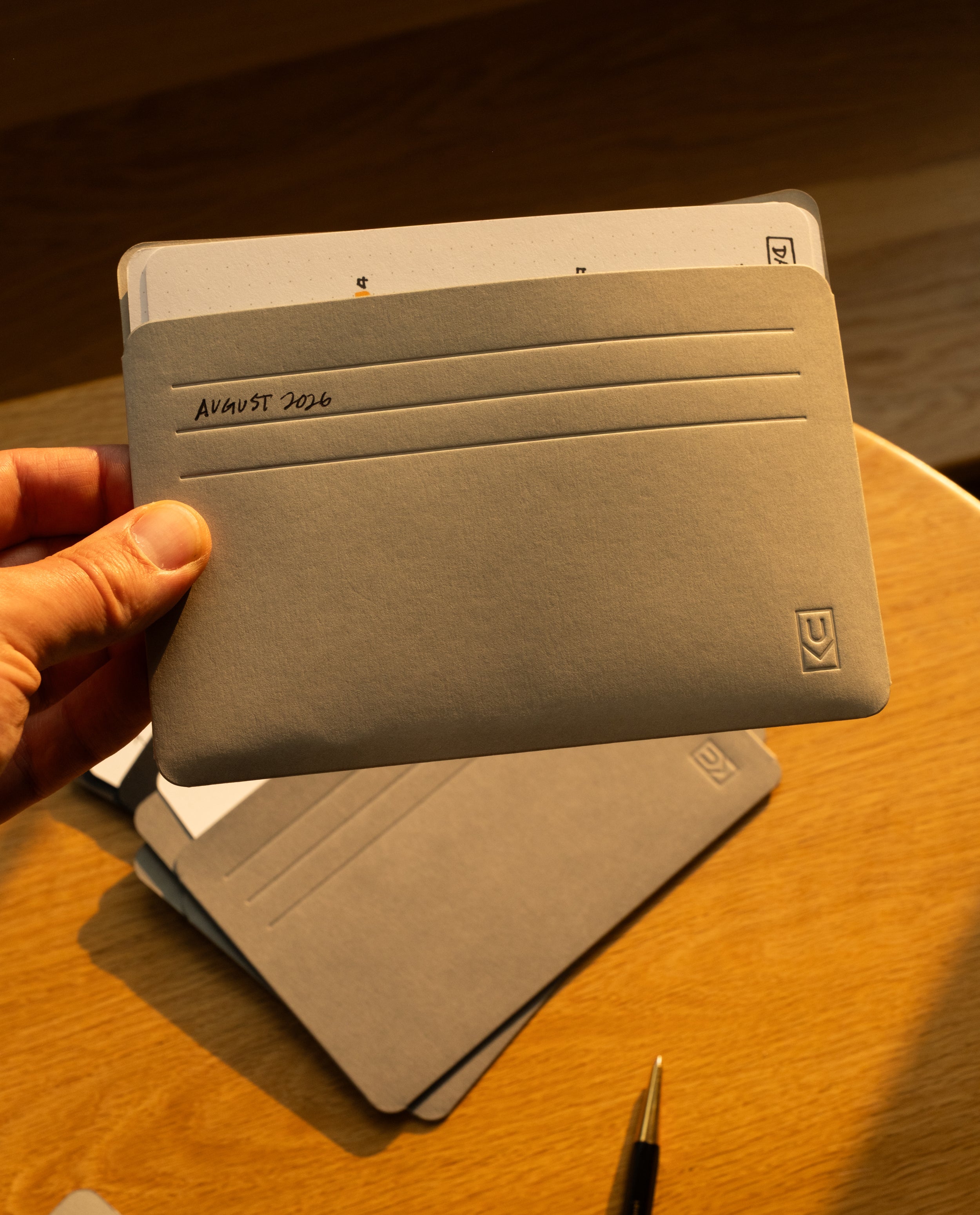 A hand holds an Analog Card Sleeve - Large (Monochromatic) in gray labeled August 2026; card organizer accessories, including additional gray Analog Card Sleeves and a pen, are arranged on a wooden surface in warm light.