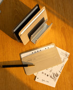 A desk organizer holds notebooks. On the wooden table, a pen rests on a tan notebook. Next to it are open note cards, neatly tucked into Analog Card Sleeves - Small (Monochrome), displaying website sketches and handwritten notes. Sunlight casts warm shadows across the scene.
