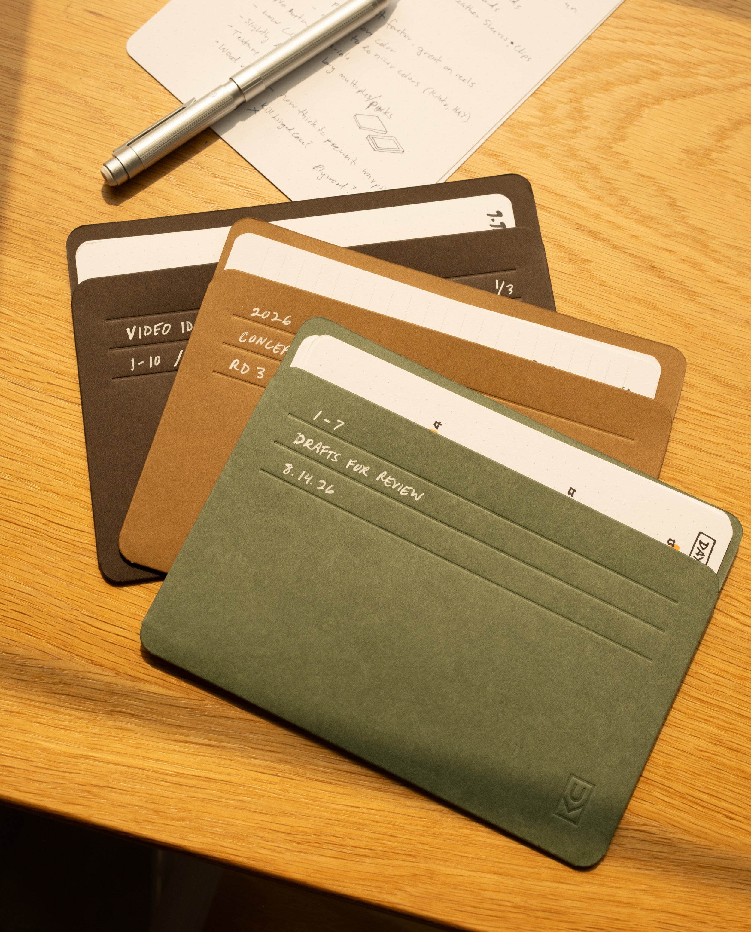 Four colored card holders—Analog Card Sleeves - Large (Earth) in green, brown, and black—are fanned out on a wooden desk near a silver pen and a sheet of notes. These holders are perfect for organizing labeled index cards, ideas, and drafts.