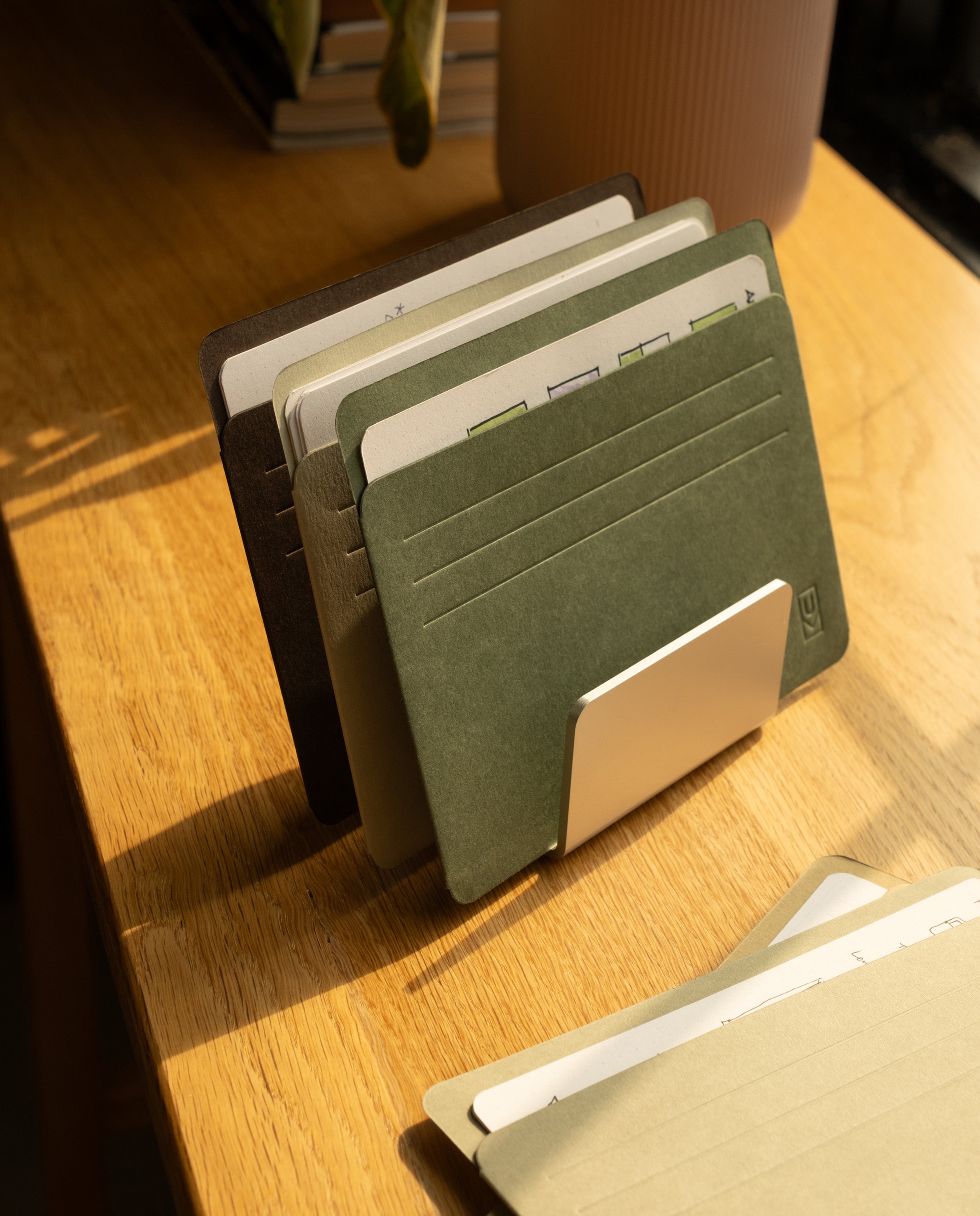 A set of file folders organized in a holder sits on a wooden desk, with sunlight streaming in. Analog Card Sleeves - Large (Earth) add a touch of order among the folders, while another folder lies flat in the foreground.