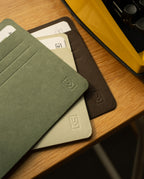 Three Analog Card Sleeves - Large (Earth)—in green, beige, and brown—rest on a wooden desk beside a yellow typewriter. Each sleeve acts as a stylish card organizer with a subtle embossed logo and slots for cards, some of which are partially visible inside. These Analog Card Sleeves - Large (Earth) blend classic design with practical function to keep your essentials neatly arranged.