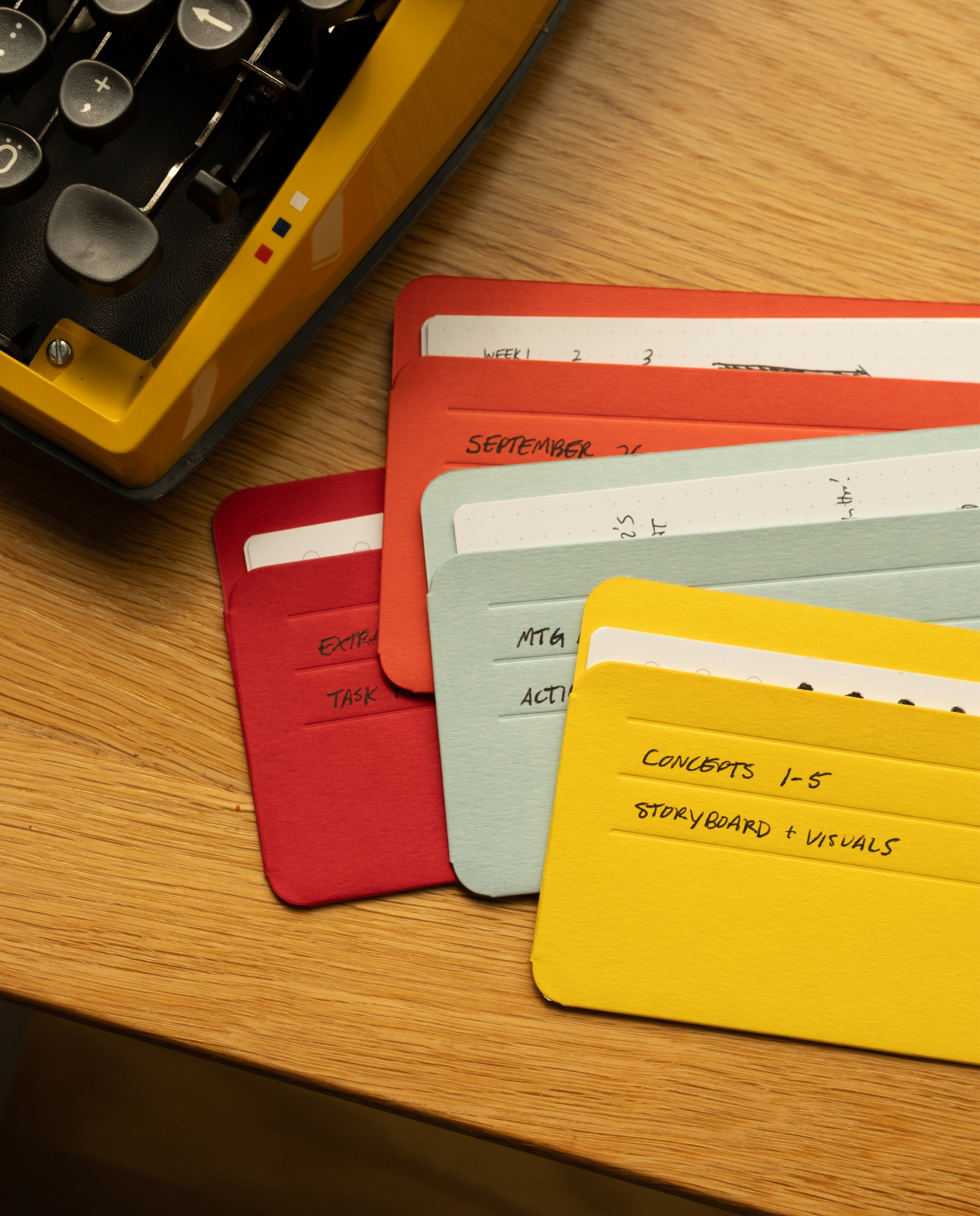 Colorful index cards with handwritten notes are spread out on a wooden desk next to the corner of a yellow typewriter. Neatly stored in Analog Card Sleeves - Small (Bright), the cards are organized by color and labeled with tasks, dates, and project concepts—a practical card organizer for any creative workspace.