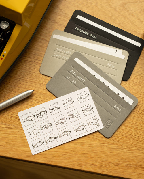 A set of monochrome cards labeled with dates and notes rests in the Analog Card Sleeves - Small (Monochrome) on a wooden surface, accompanied by a silver pen and a yellow object. One card features small sketches arranged in a tidy grid, all kept organized and accessible in this practical card organizer.