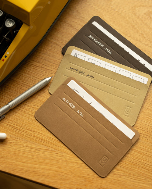 Three labeled monthly planner cards—October, September, and November 2024—are spread on a wooden desk next to a silver pen and a yellow typewriter. Each card fits neatly into your card organizer or Analog Card Sleeves - Small (Earth), with white spaces for notes or schedules.