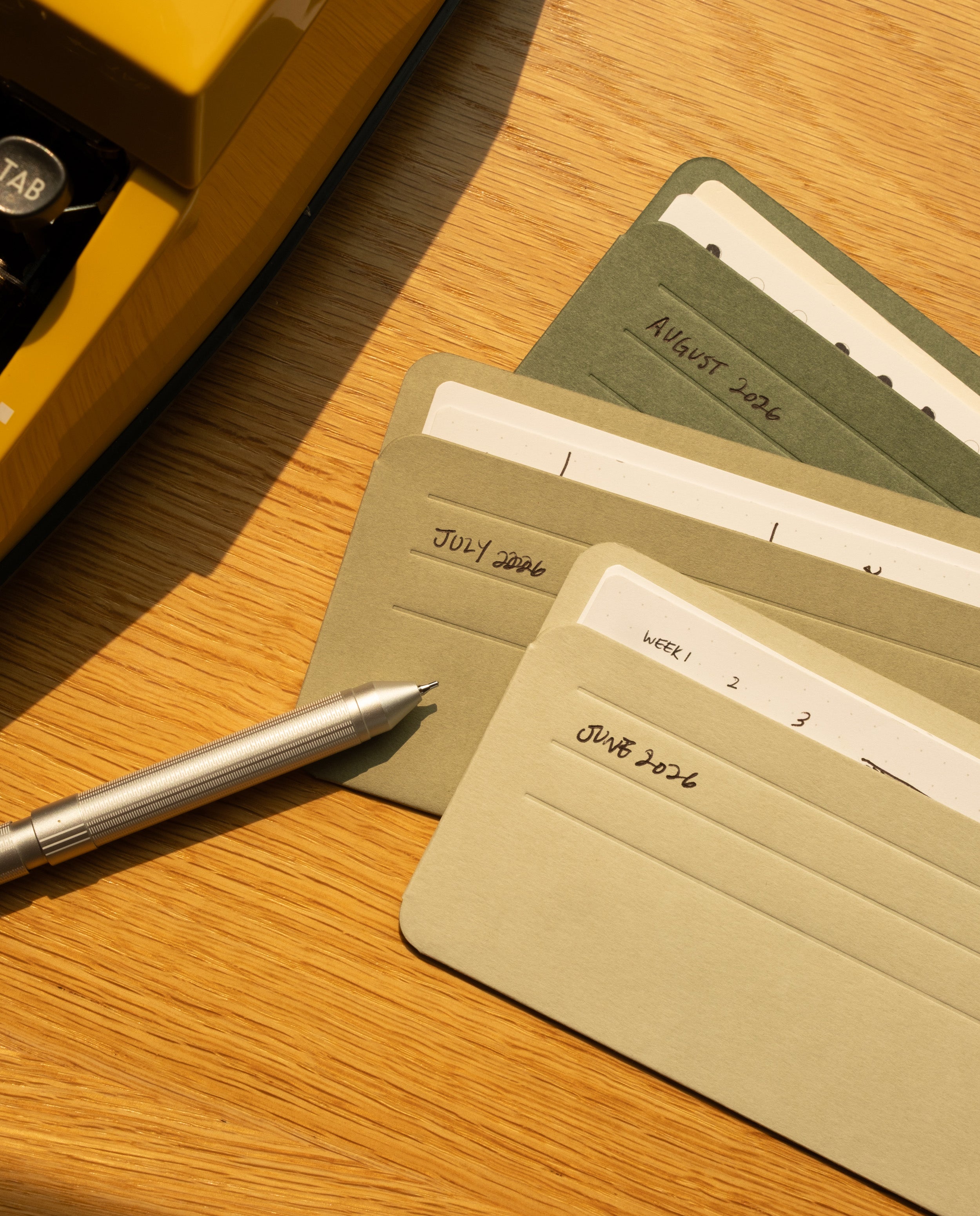 A silver pen rests on a wooden desk next to green and beige index cards labeled June 2026, July 2026, and August 2026, neatly arranged in an Analog Card Sleeves - Small (Earth) card organizer. Part of a yellow typewriter is visible in the top left corner.
