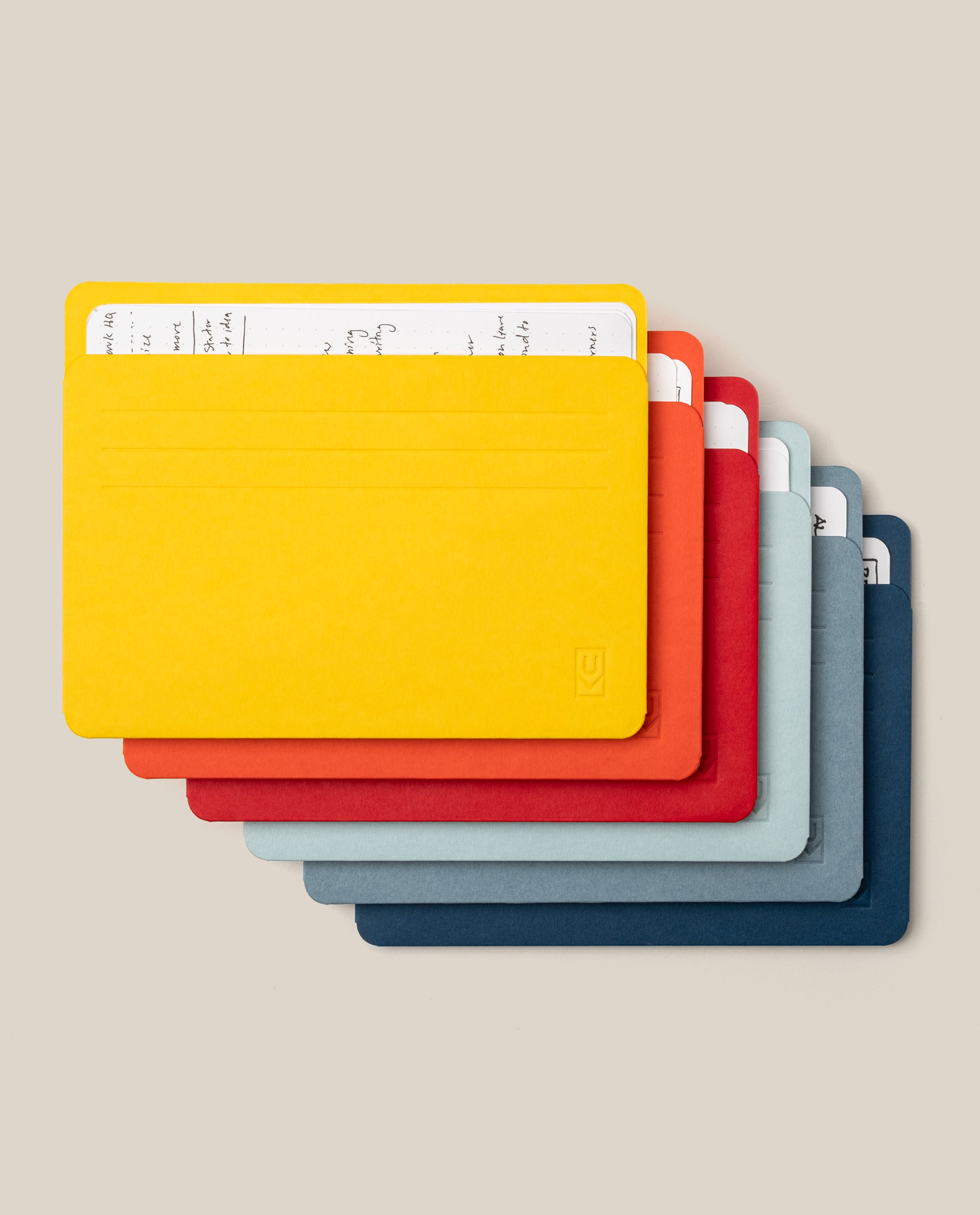 Six brightly colored Large Analog Card Sleeves (Set of 6 - Bright)—in yellow, orange, red, dark gray, light gray, and blue—are stacked together with a handwritten note partially visible in the top yellow sleeve. Ideal for anyone seeking to organize cards efficiently, these vibrant sleeves stand out against a neutral beige background.