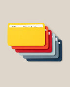 Six minimalist Analog Card Sleeves - Small (Bright) in yellow, orange, red, light blue, navy blue, and grey are stacked in a fan arrangement on a beige background, serving as a stylish card organizer with cards partially visible in the top holder.