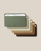 Six minimalist Analog Card Sleeves - Small (Earth) in green, light gray, tan, light brown, brown, and dark brown are stacked and fanned out as a stylish card organizer. Each sleeve holds white cards and is displayed on a beige background.