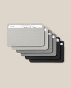 Six Analog Card Sleeves - Small (Monochrome) in grey and black overlap in a fan shape on a beige background, each featuring visible horizontal lines and rounded corners. Ideal for your card organizer or storage system, the top sleeve displays a partial handwritten note.