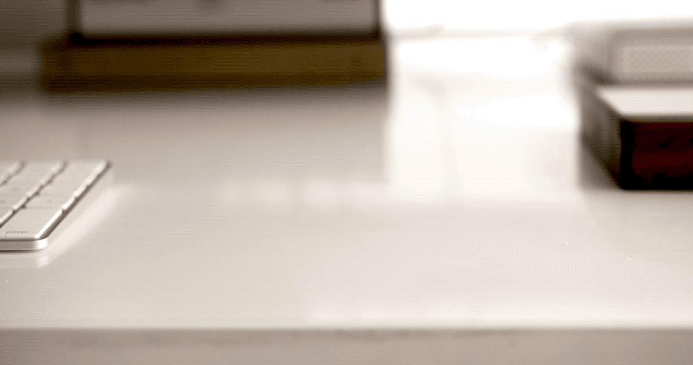 A close-up of a white desk with a wireless keyboard on the left and a blurred box or book on the right, with soft lighting creating shadows and a minimal, tidy workspace feel.