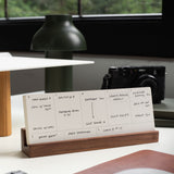 An Analog Weekly Card from our kit is displayed in a wooden holder on a desk, organized with tasks and appointments. A camera and greenery are blurred in the background. Text reads: "*Card Holder sold separately.