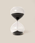The 15 Minute Hourglass (Clear), featuring black sand with half already settled in the lower chamber, stands against a plain beige background—an elegant analog timer that beautifully marks the quiet passage of time.