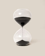 The 15 Minute Hourglass (Clear), featuring black sand with half already settled in the lower chamber, stands against a plain beige background—an elegant analog timer that beautifully marks the quiet passage of time.