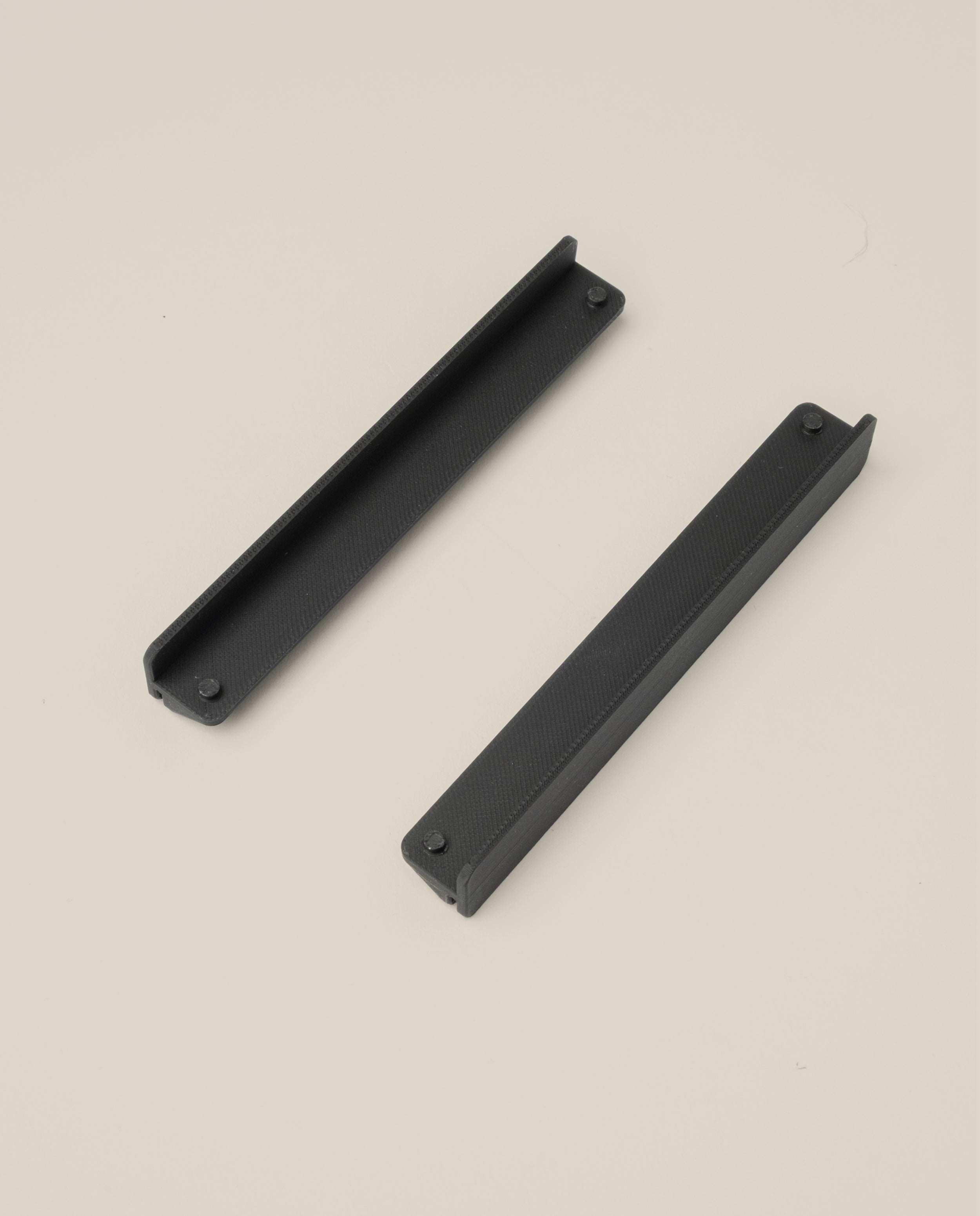 Two black, rectangular Warehouse Item - Capture Tray Clips (Black - Large) with textured surfaces are positioned diagonally apart on a plain light beige background, showcasing their robust design.