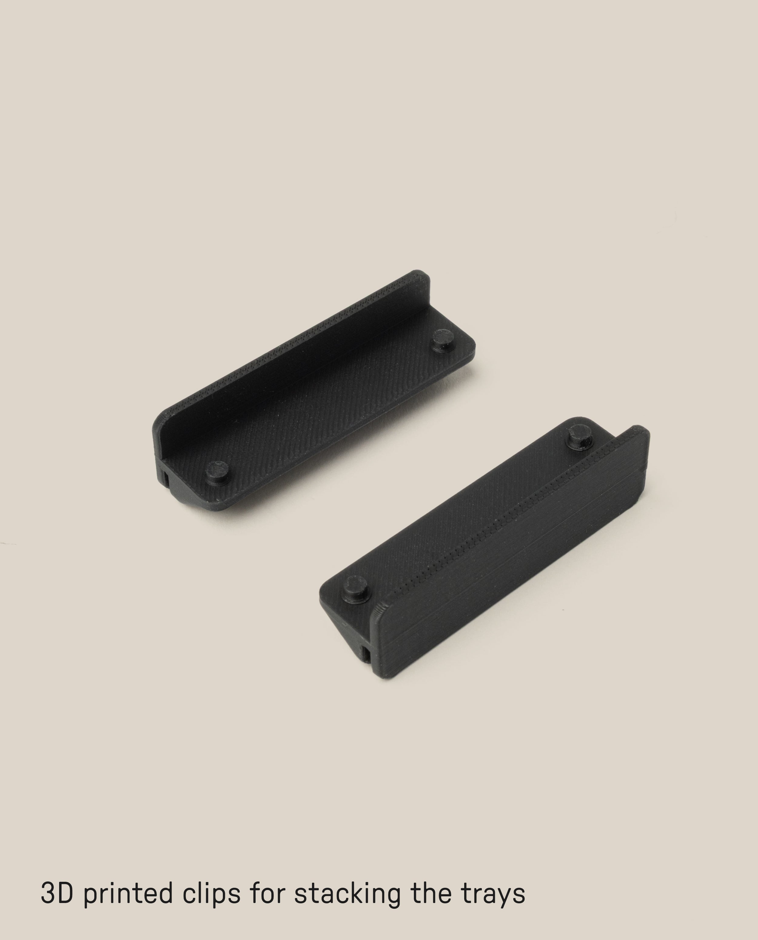 Two black 3D printed clips are displayed on a beige background. Designed for the Capture Bundle (Small Tray + Large Tray - Black Steel), the rectangular clips have raised edges to securely stack the trays. Text: 3D printed clips for stacking the trays.