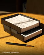 Two black paper trays are stacked on a wooden desk, each containing a notepad filled with sketches and notes. The Warehouse Item - Capture Tray Clips (Black - Large) secure the trays together, providing effortless organization. Nearby rests a black and gold pen. Sunlight streams in, casting shadows across the scene. In the corner, text reads “*Trays sold separately.”.