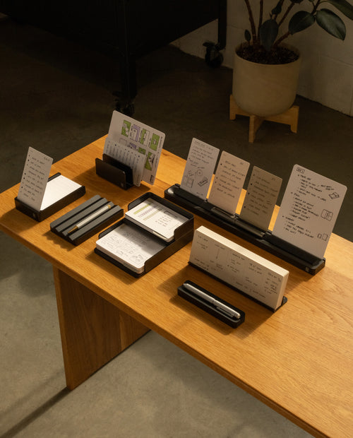 A wooden table displays The Complete Analog Collection (Black Aluminum) desktop organizer, holding note cards, notebooks, pens, and papers with handwritten notes and sketches. A potted plant rests in the background on a concrete floor.