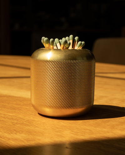 The Craighill Match Striker (Brass), crafted from golden, textured solid brass, holds dozens of green-tipped strike anywhere matches and rests elegantly on a sunlit wooden surface.