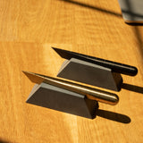 A gold and a black metallic pen rest diagonally on Craighill Desk Knife Plinths (Slate), triangular cast concrete holders arranged on a wooden surface, with sunlight casting shadows across the scene.