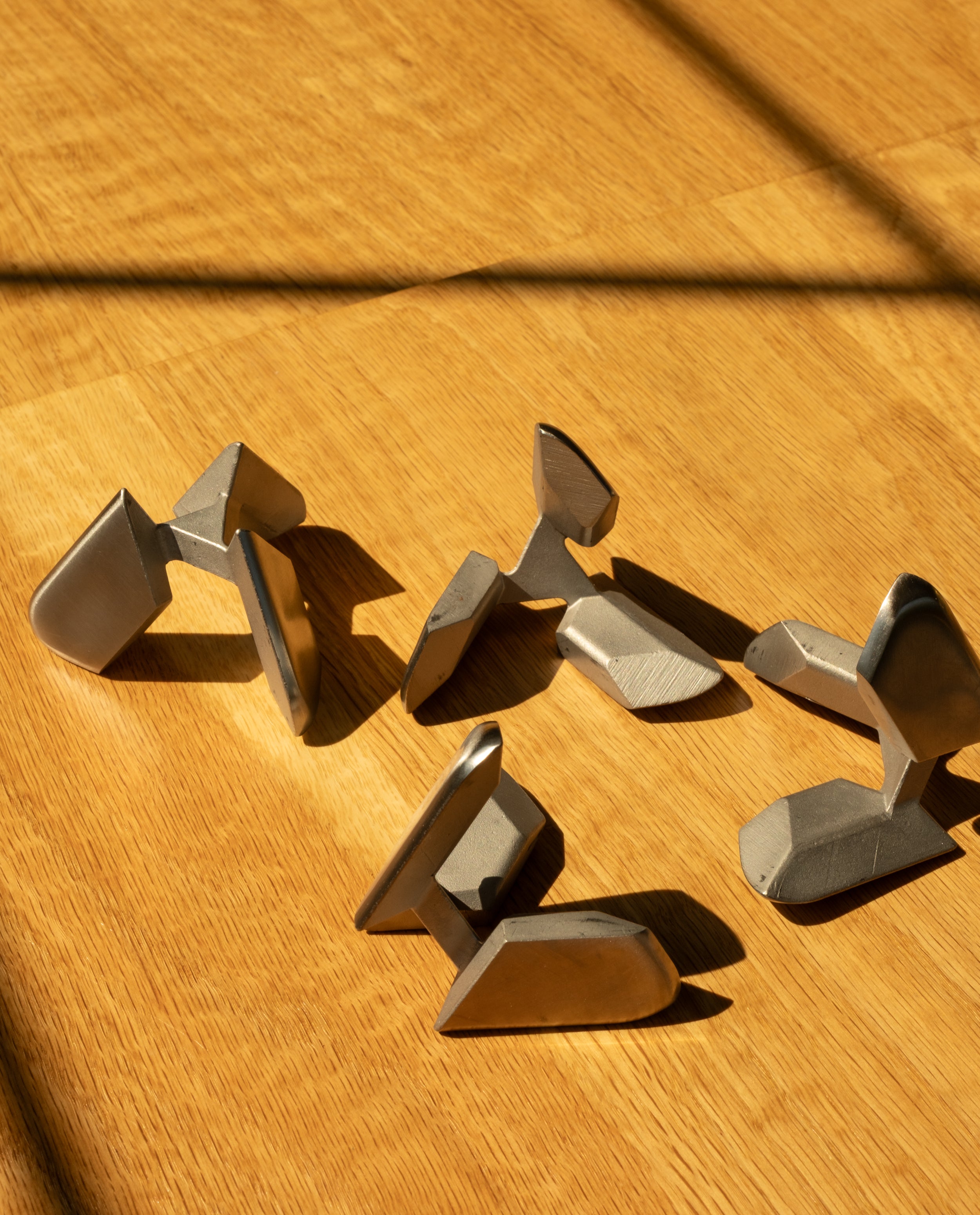 Six abstract metal sculptures rest on a wooden surface, sunlight casting angled shadows across them—each form reminiscent of the Craighill Tetra Puzzle in stainless steel, elegantly blending artistry with mechanical intrigue.