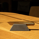 A Craighill Desk Knife with a gold handle rests on a Craighill Desk Knife Plinth (Slate), both positioned on a wooden table where sunlight casts shadows across the surface.