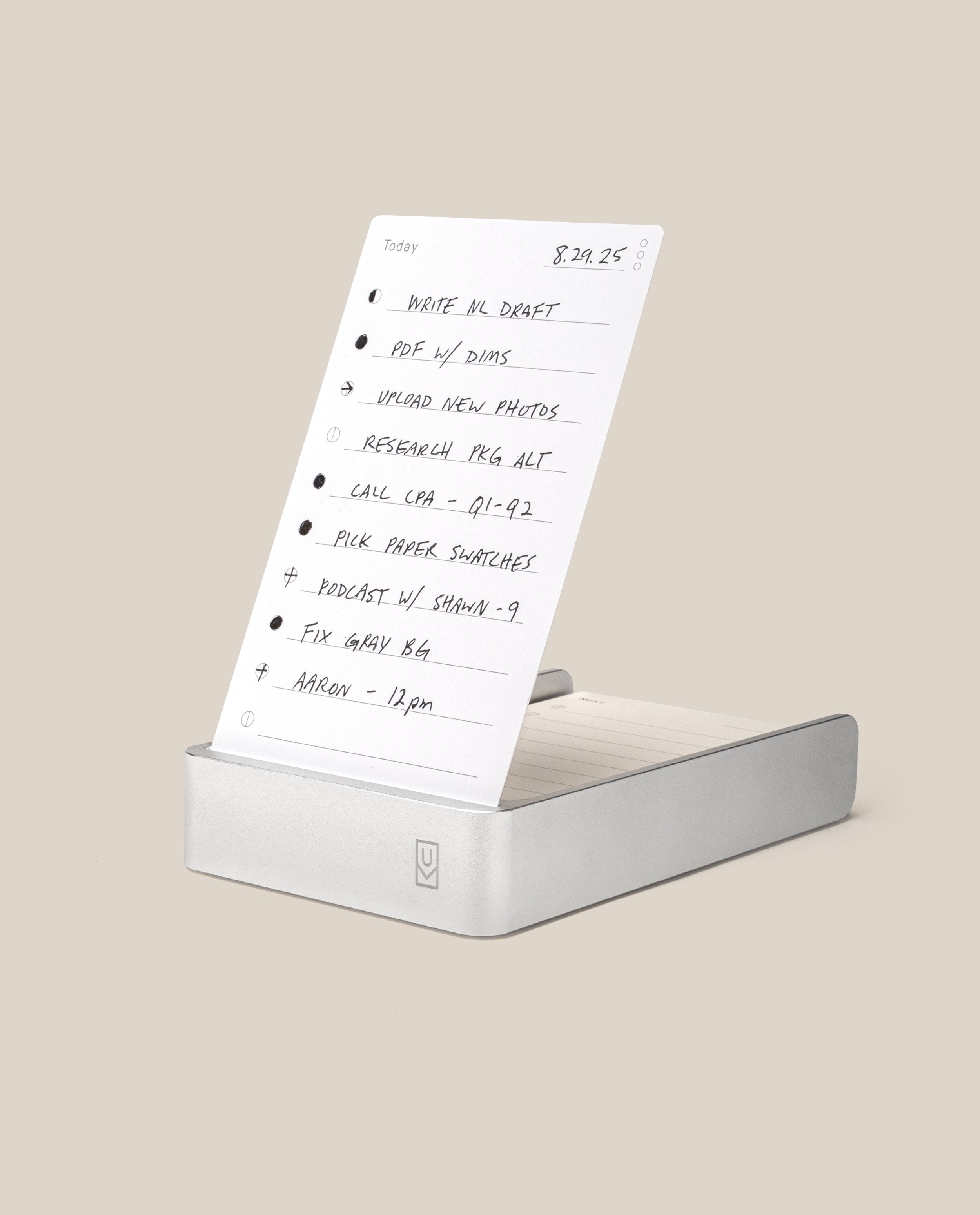 A handwritten to-do list on white paper stands upright in the Analog Daily Focus Kit (Silver Aluminum). Tasks like writing, photo uploads, research, and meetings are neatly penned in black ink. Background is light beige.