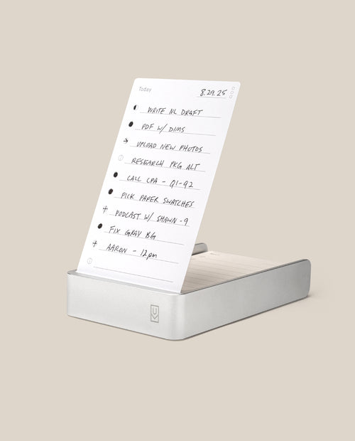 A handwritten to-do list on white paper stands upright in the Analog Daily Focus Kit (Silver Aluminum). Tasks like writing, photo uploads, research, and meetings are neatly penned in black ink. Background is light beige.