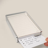 A hand pulls a task card from the Analog Daily Focus Kit + 6 Refills (Silver Aluminum), revealing a handwritten to-do list on a lined card—an elegant analog productivity system against a clean, light-colored background.