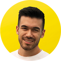 A man with short dark hair and a trimmed beard smiles at the camera, standing in front of a bright yellow circular background. He is wearing a white shirt.