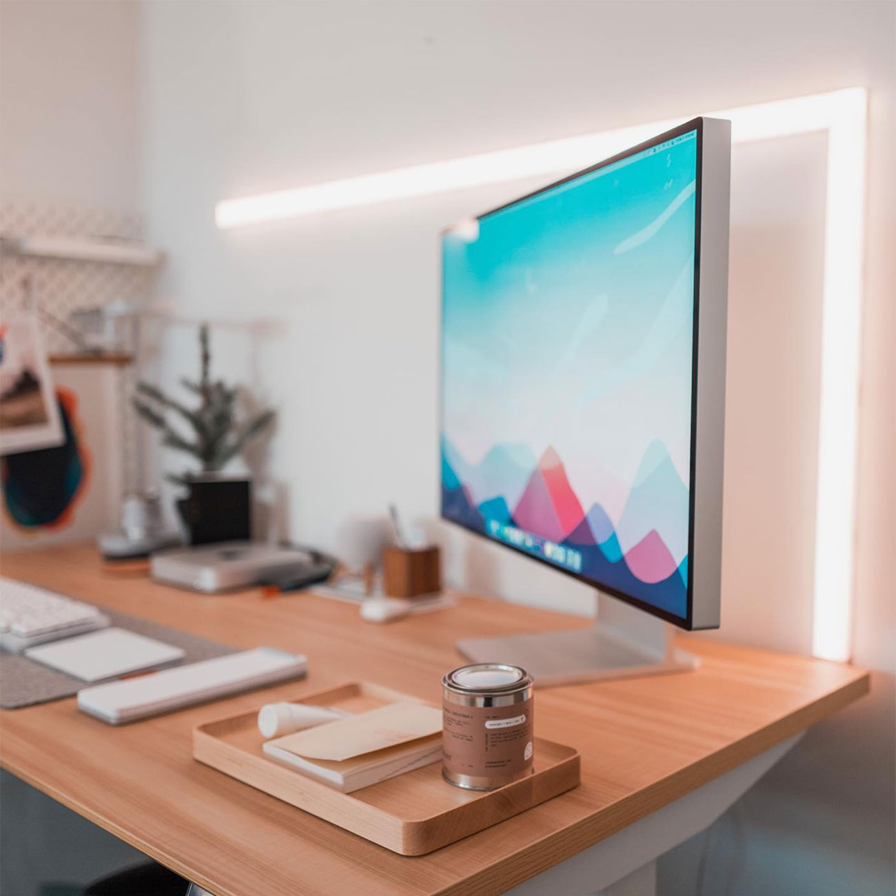A modern, minimal desk setup with a large monitor displaying a colorful abstract wallpaper, a wooden tray with a candle and notepad, a keyboard, and a small potted plant, all on a light wooden desk.