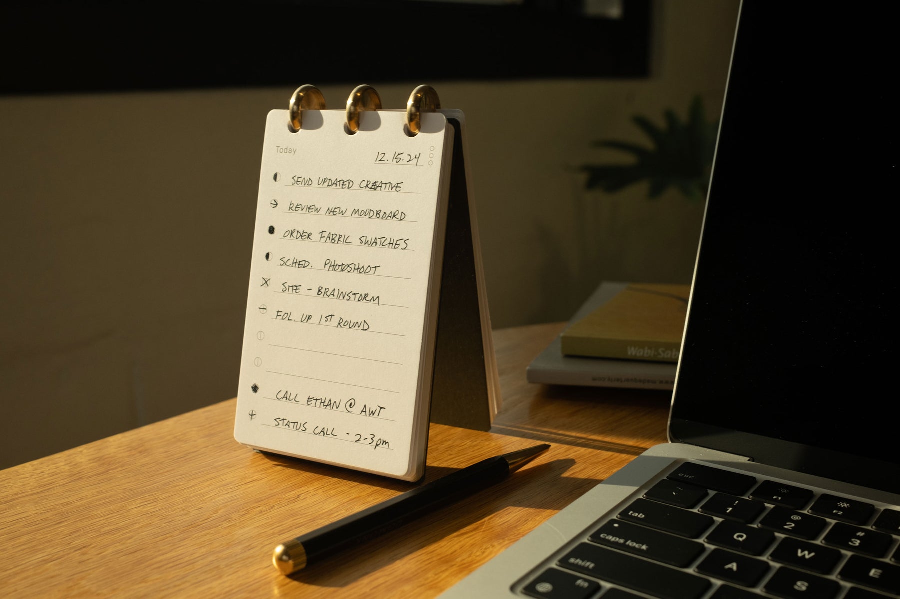A notepad with a to-do list stands on a wooden desk next to a laptop. The list includes tasks like sending updates, ordering samples, and calls. Warm sunlight illuminates the scene.