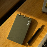 A Discbound Analog + 3 Refills notepad with brass disc binding rests on a wooden desk beside a black and gold pen and the corner of a laptop keyboard.