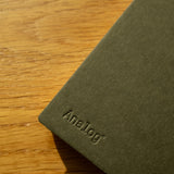 A Discbound Analog + 3 Refills notebook with brass disc binding rests on a wooden surface, soft sunlight highlighting the dark green cover and textured materials.