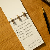 The Discbound Analog + 3 Refills, a compact notepad with brass discs, sits on a wooden desk beside a black and gold pen, open to a handwritten to-do list for creative tasks, fabric swatches, photoshoots, and calls.