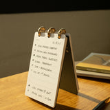 The Discbound Analog + 3 Refills, a compact refillable planner for handwritten tasks, to-do lists, and scheduled calls with brass disc binding, rests on a wooden table beside a pen and book in warm sunlight.