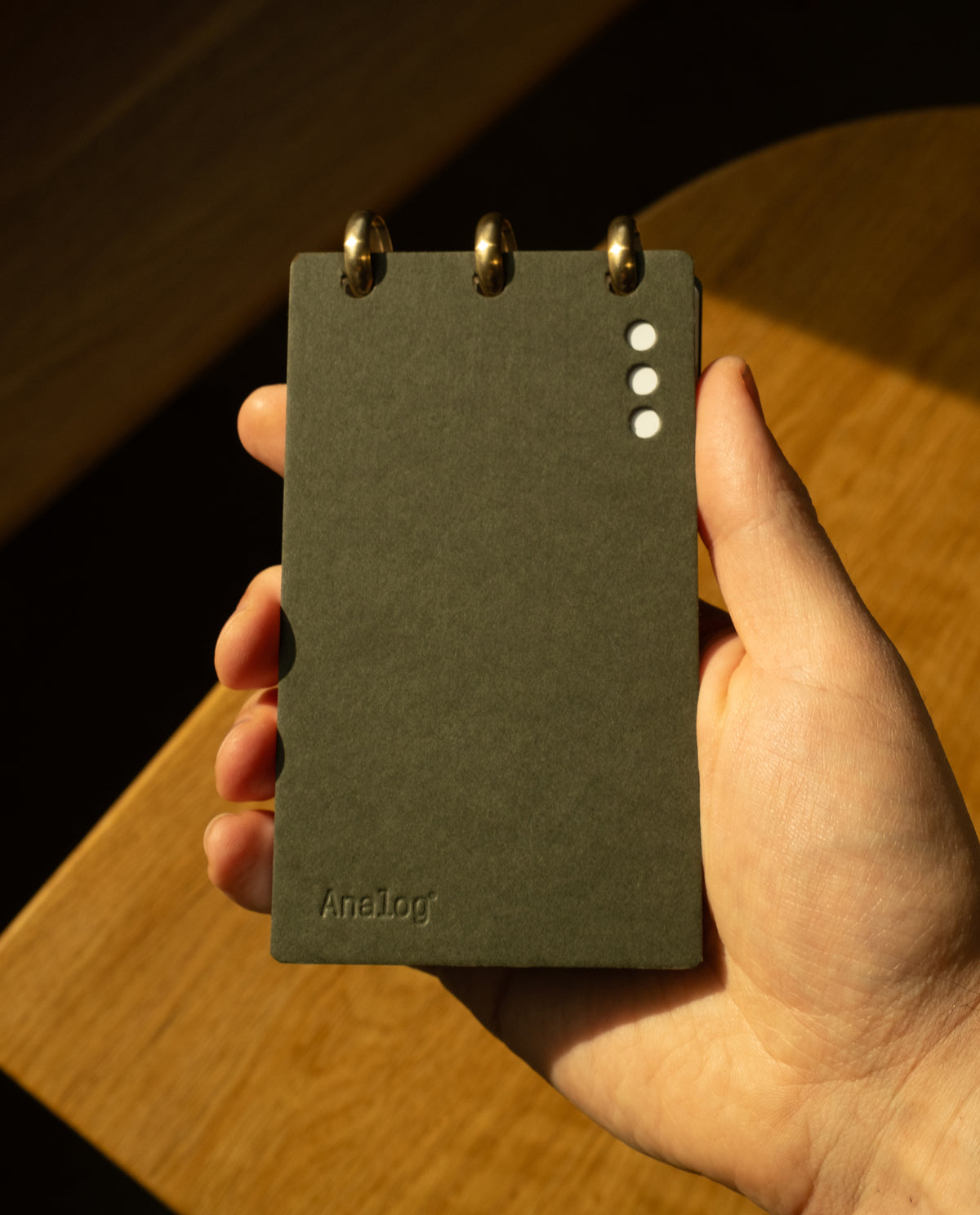 A hand holds a small, dark gray Discbound Analog + 3 Refills notepad with brass disc binding at the top. The cover has three white dots and 