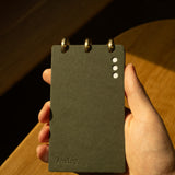 A hand holds a small, dark gray Discbound Analog + 3 Refills notepad with brass disc binding at the top. The cover has three white dots and "Analog" embossed, lit warmly with soft shadows on the hand and wooden surface.