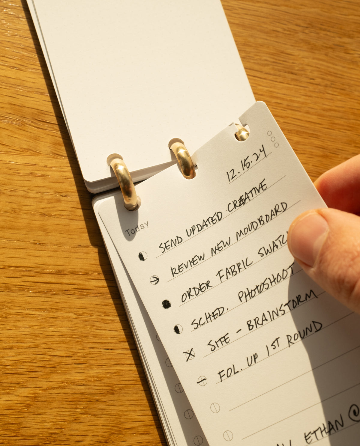 A hand holds a small ring-bound notepad on a wooden surface. The visible page lists tasks including Send updated creative, Review new moodboard, and more, with some items checked or crossed out.