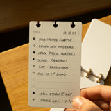 A hand holds the Discbound Analog + 3 Refills planner with brass disc binding, open to a to-do list for 12.15.24: send creative updates, review a moodboard, order fabric swatches, make calls—bathed in warm sunlight.