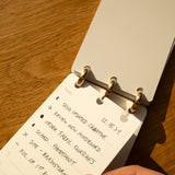 Close-up of a hand holding a Discbound Analog + 3 Refills planner on wood, displaying a handwritten to-do list for 12-16-24 with tasks like creative work, moodboard, fabric swatches, and photoshoot.