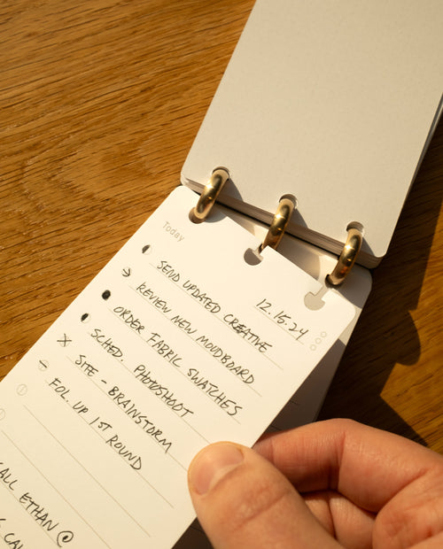 A hand holds an open Discbound Analog notepad displaying a to-do list with tasks like sending updated creative, removing an old moodboard, and scheduling a photoshoot. Warm sunlight highlights the wooden surface below, while a Discbound Analog Refill (6-pack) is stacked neatly nearby.
