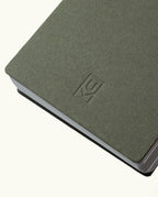 A close-up of the Discbound Analog + 3 Refills notebook in gray-green, featuring a simple embossed KC logo, brass disc binding, rounded corners, and slightly fanned pages against a plain background.