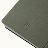 A close-up of the Discbound Analog + 3 Refills notebook in gray-green, featuring a simple embossed KC logo, brass disc binding, rounded corners, and slightly fanned pages against a plain background.