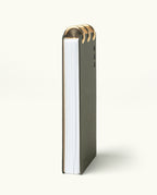 The Discbound Analog + 3 Refills is a vertically standing notebook with a dark cover and thick white pages, bound at the top with three brass disc binding rings, shown on a plain light background.